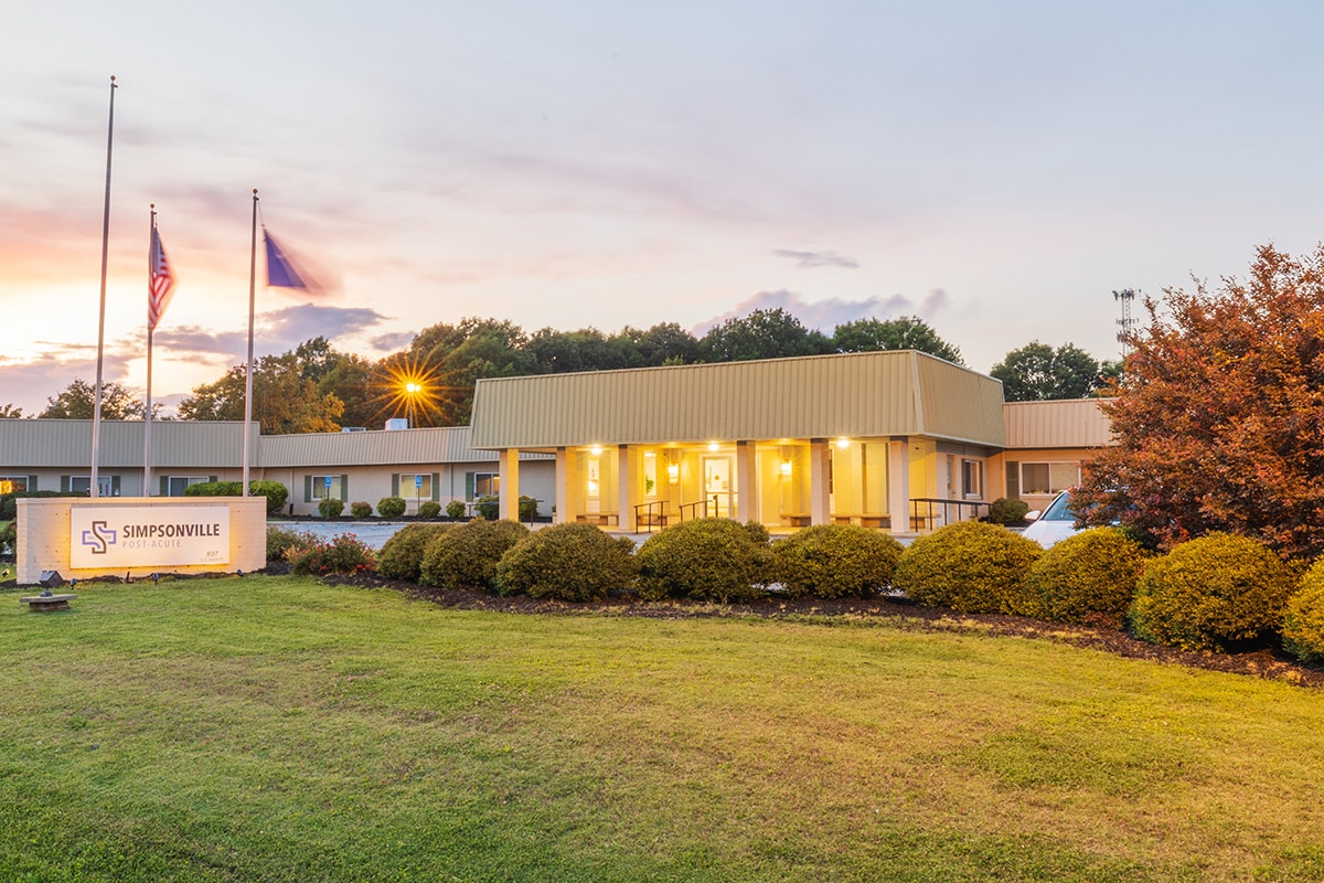 Exterior view of Simpsonville Post Acute at dusk with signage and American flag flying out front.